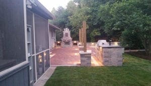 A backyard patio with a grill and a fireplace. The patio is surrounded by trees.